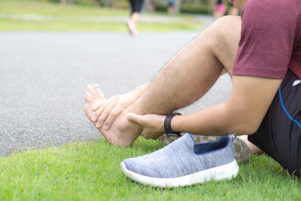 Athletic man sitting on grass beside a running path holding his painful heel and ankle after exercise, highlighting need for professional care from Foot Doctor Titusville for foot and heel pain relief